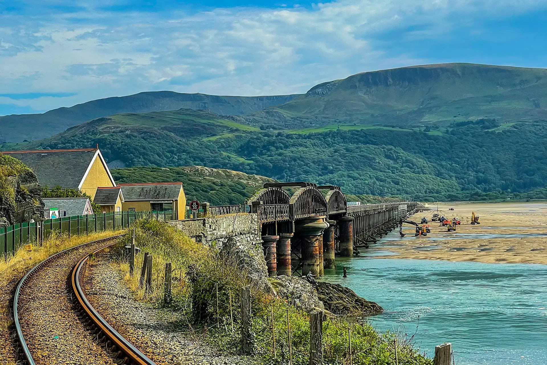 A train line over a river bank with construction in the foreground, with British countryside landscape in the background.