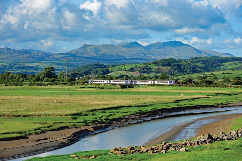 A train travelling in Cumbria, with a river in the foreground, and hills in the background.