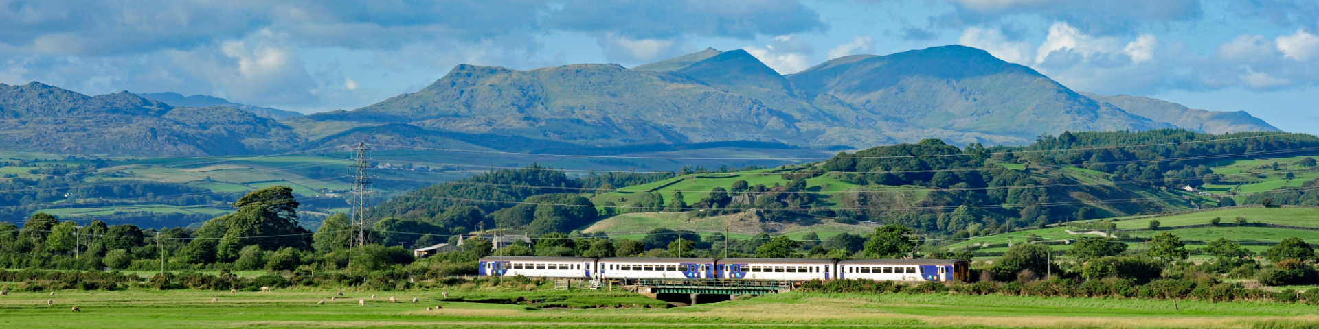 Green countryside with water, beyond a train travels in front of mountain peaks with blue sky