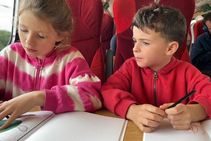 Two children sat on a train doing paper-based activities