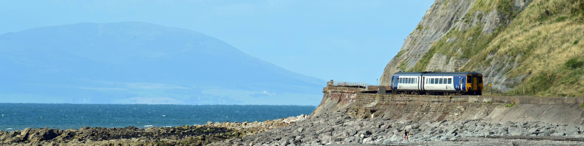 Train travelling by the side of a rocky coast with water and mountains