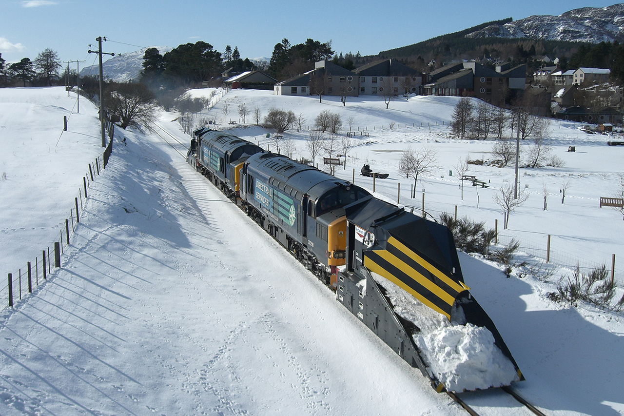 Train with snow scoop on the front clearing snow from the tracks. Snowy hills and countryside surround