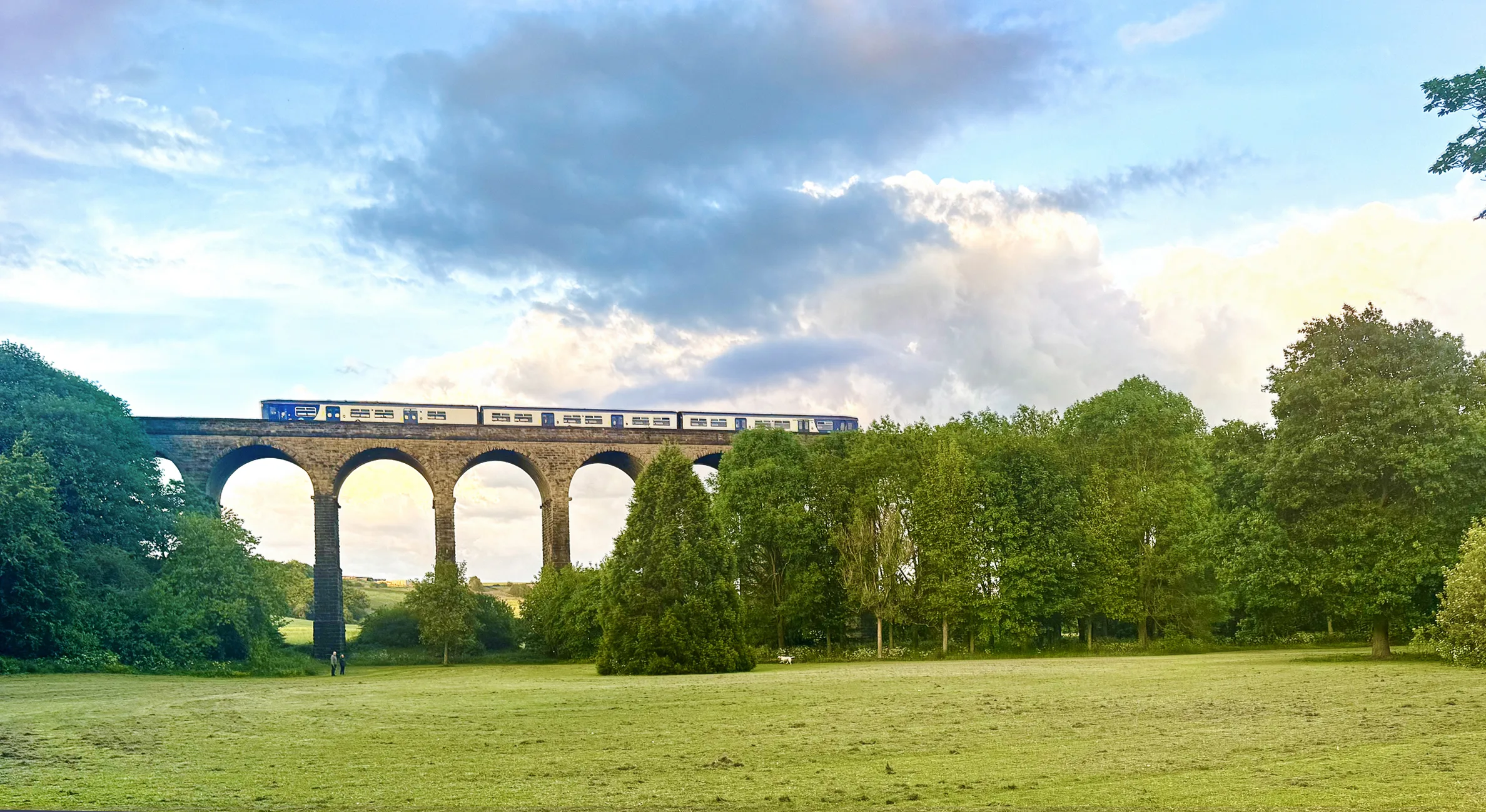 Train travelling across Penistone Viaduct with lush green trees and countryside surrounding.