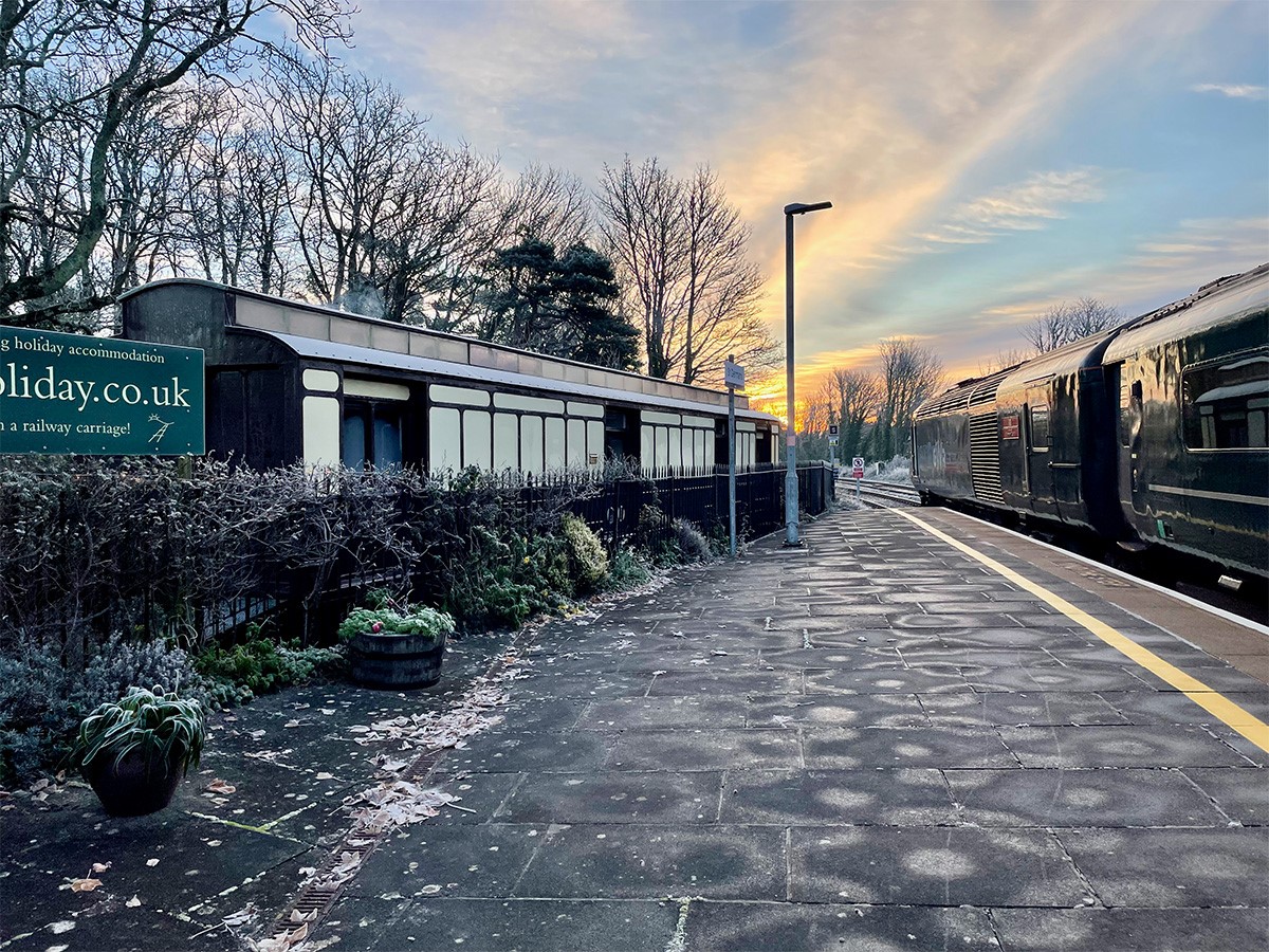 St Germans station in the winter sun with a train at the platform and a view of the rail holiday carriage on the left