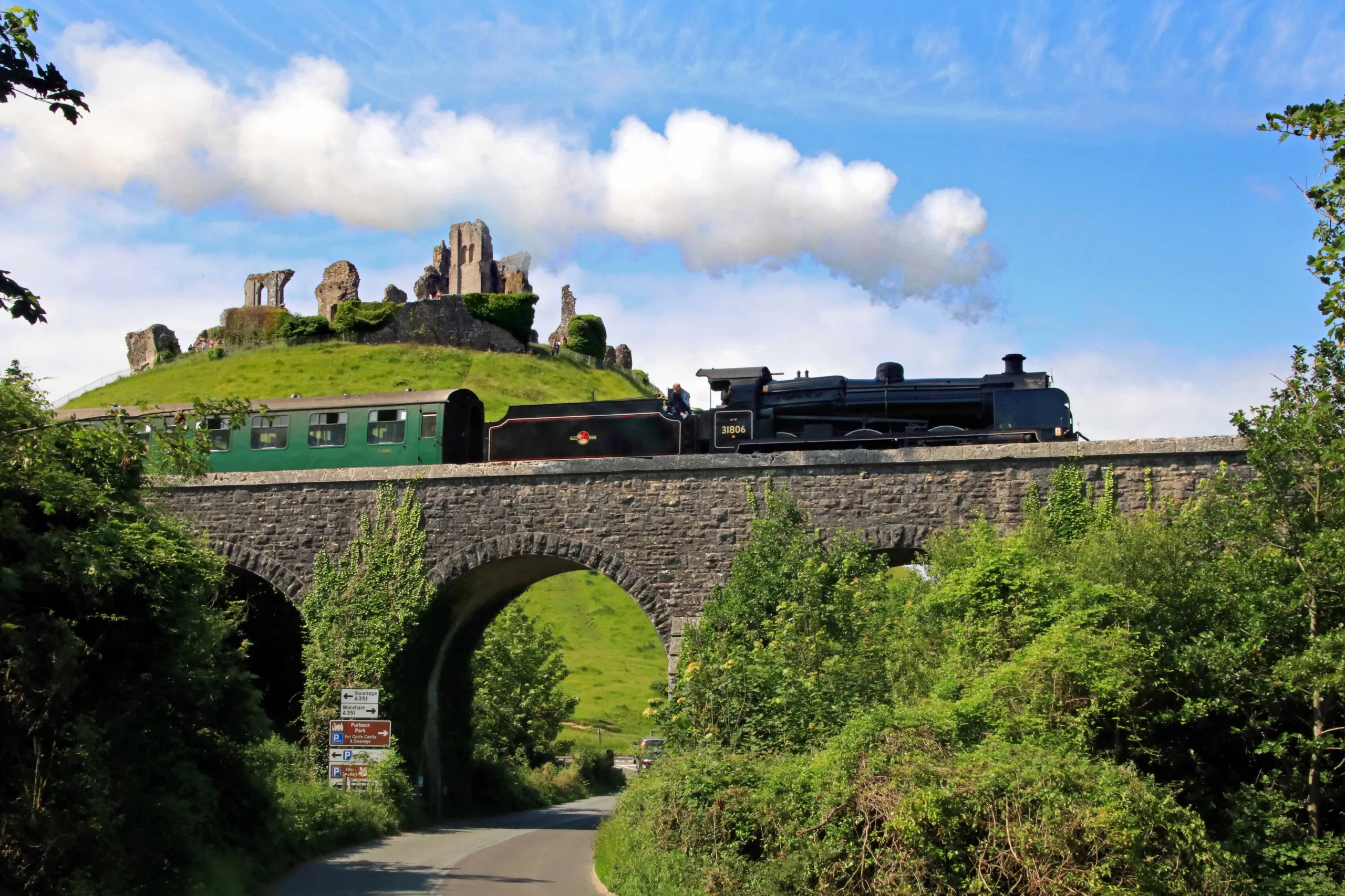 Steam train crossing over a small viaduct with historic ruins of castle on the top of a mound, in the background. White steam comes from the train and is a bright contrast against the blue sky. Purbeck Line Swanage Railway.