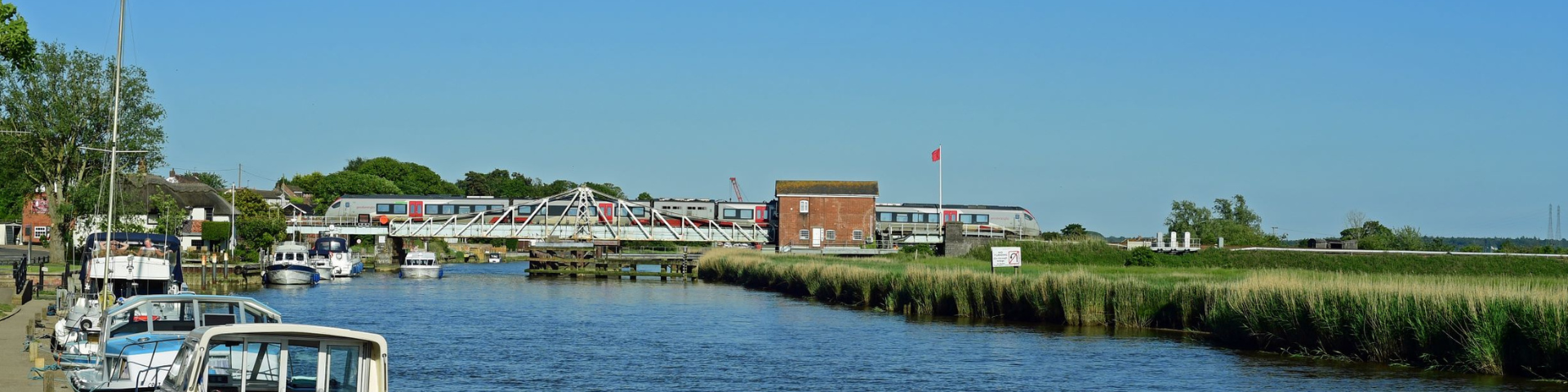 Wherry Lines train travelling across canal with boats moored up with blue sky