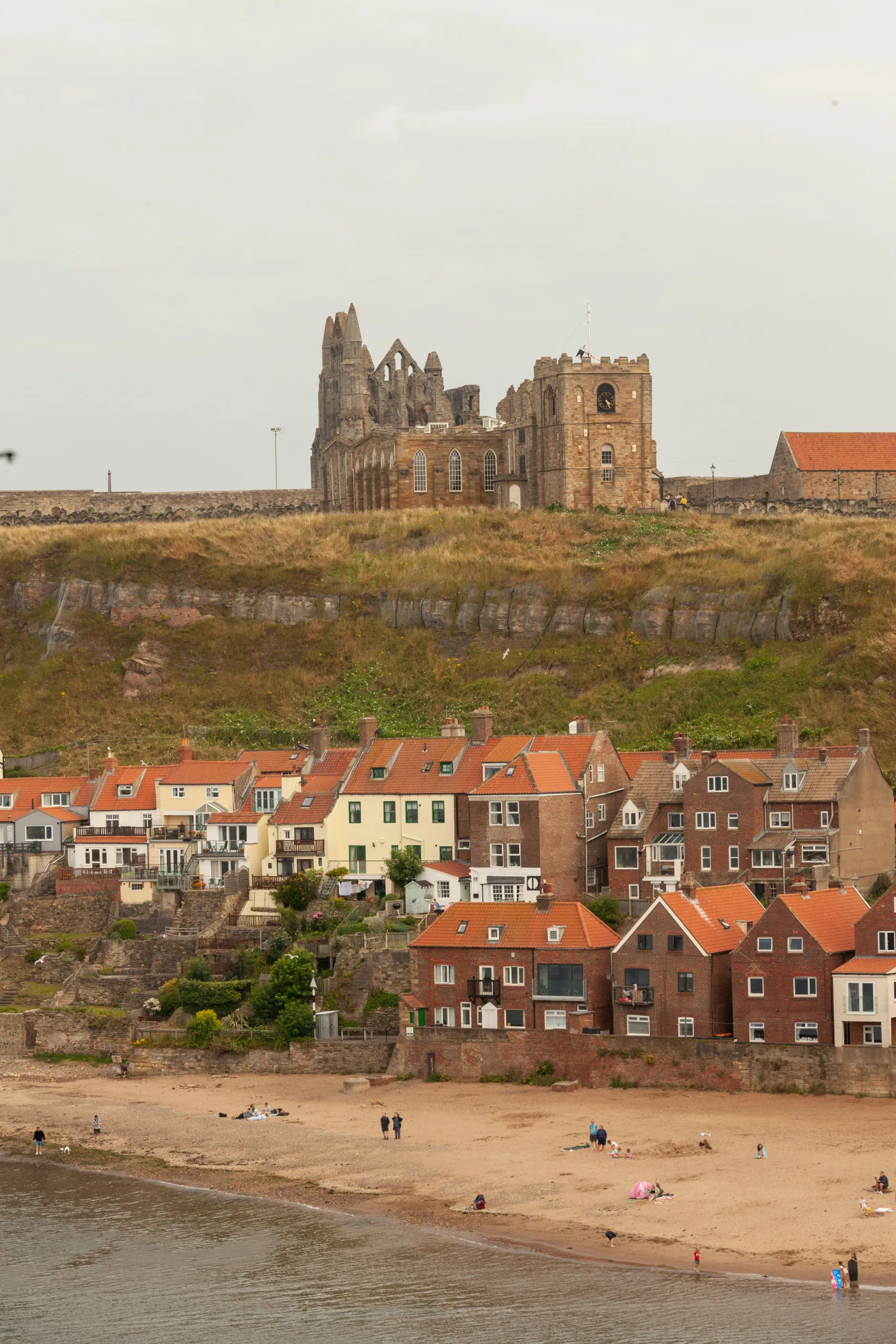 Ruins of Whitby Abbey on the top of hill with terrace houses hugging the coast by the beach below | literary trails and tails by rail