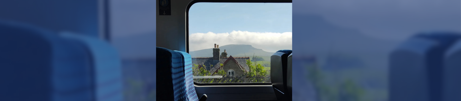 Framed by a train seat and window, this view shows a stone built house overlooked by a mountain with it's top covered by cloud and above that blue sky #scenicrailview