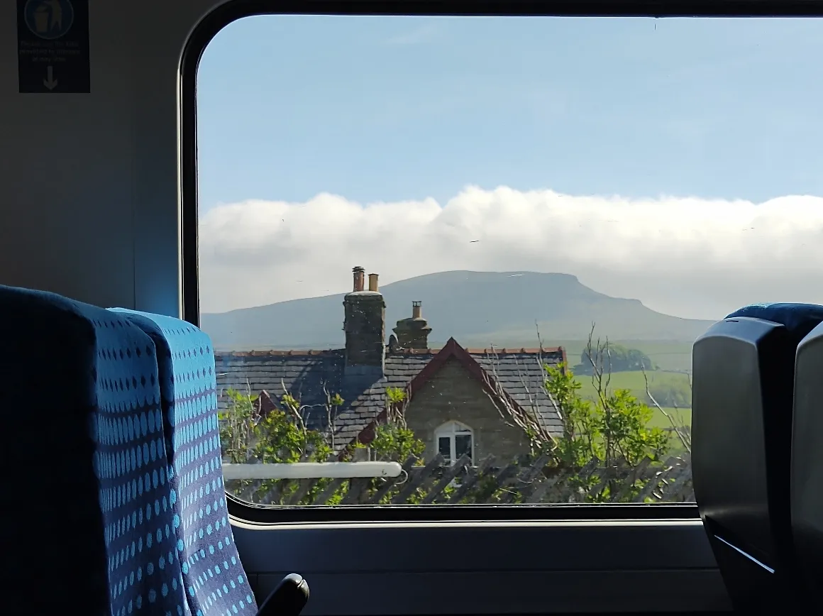 Framed by a train seat and window, this view shows a stone built house overlooked by a mountain with it's top covered by cloud and above that blue sky #scenicrailview