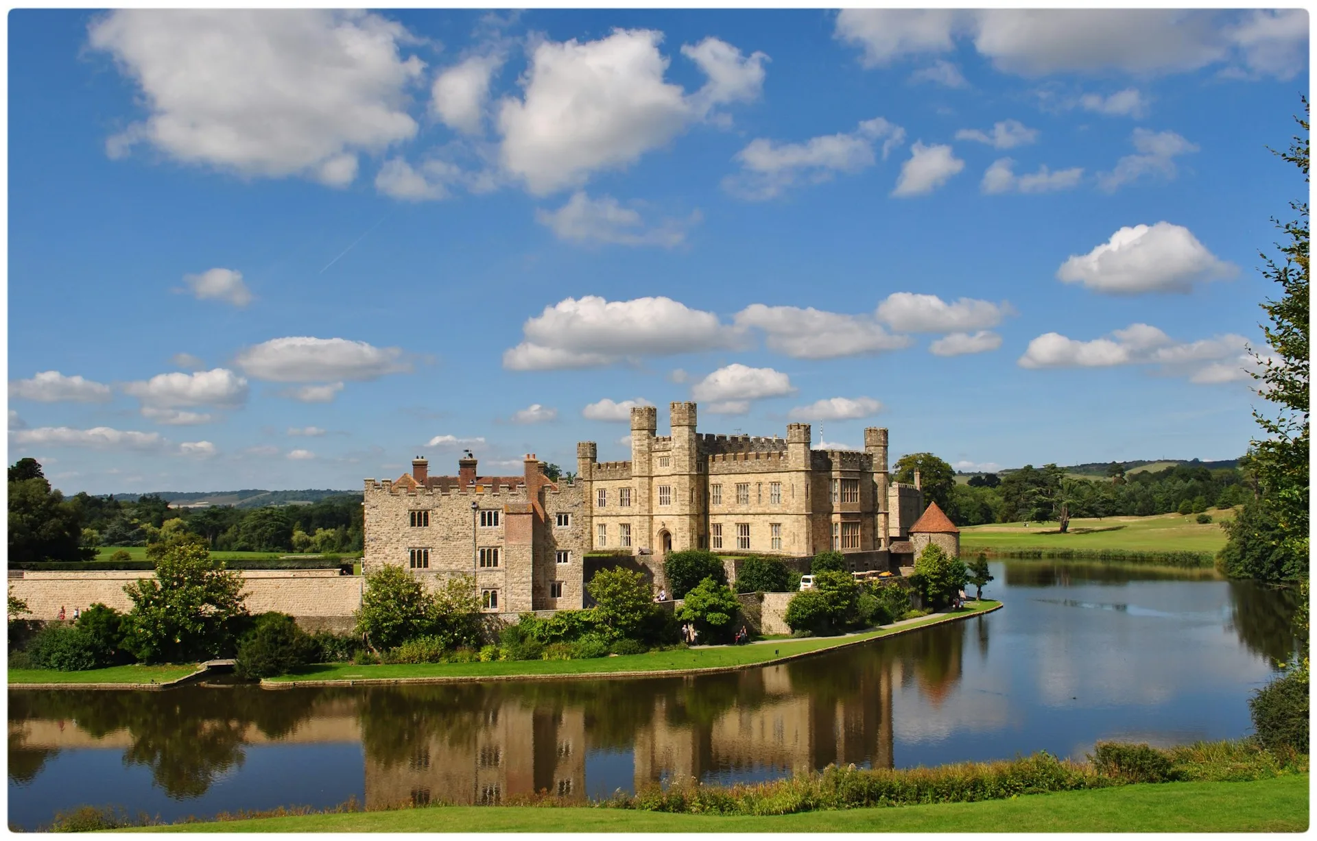 Magestic castle sits at the side of a body of water reflecting the blue sky.