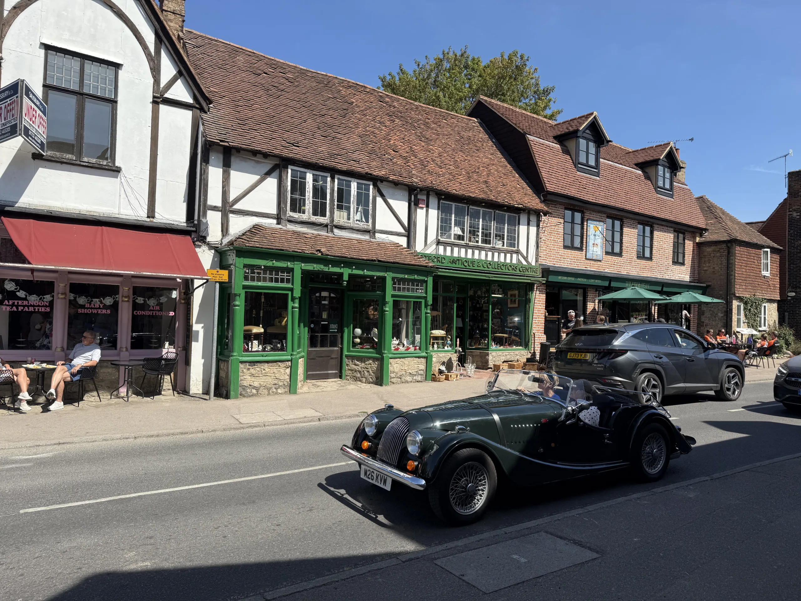 Historic buildings and independent shops on Otford High Street in Kent, with outdoor café seating and a classic green vintage car driving past on a sunny day.