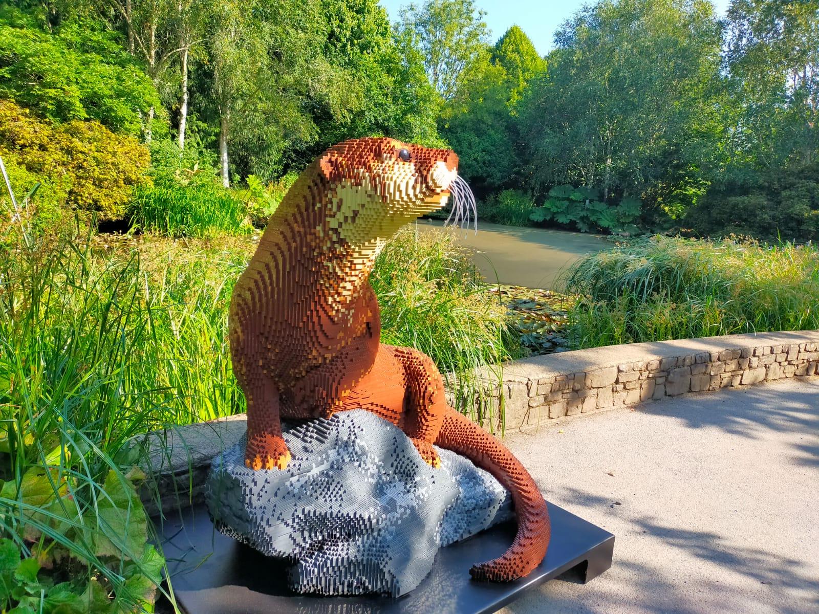 An otter made from Lego bricks sits among lush green foliage with a body of water in the background on a sunny day. Part of the 'enjoy discounts and special offers this summer' blog.