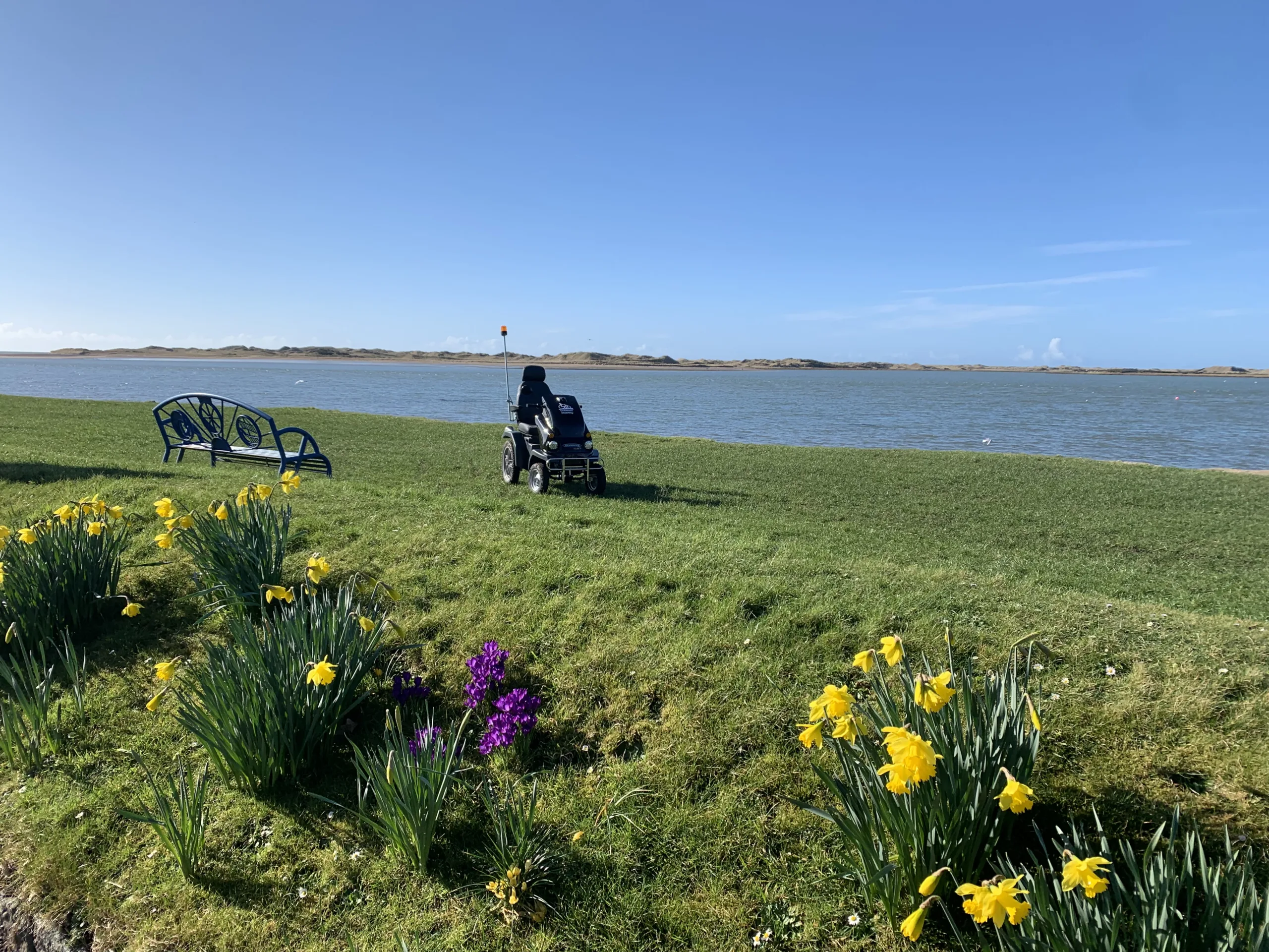 Mobility scooter parked on grass by some daffodils with a peaceful body of water in the backgroun on a blue sky day. Making amazing places accessible