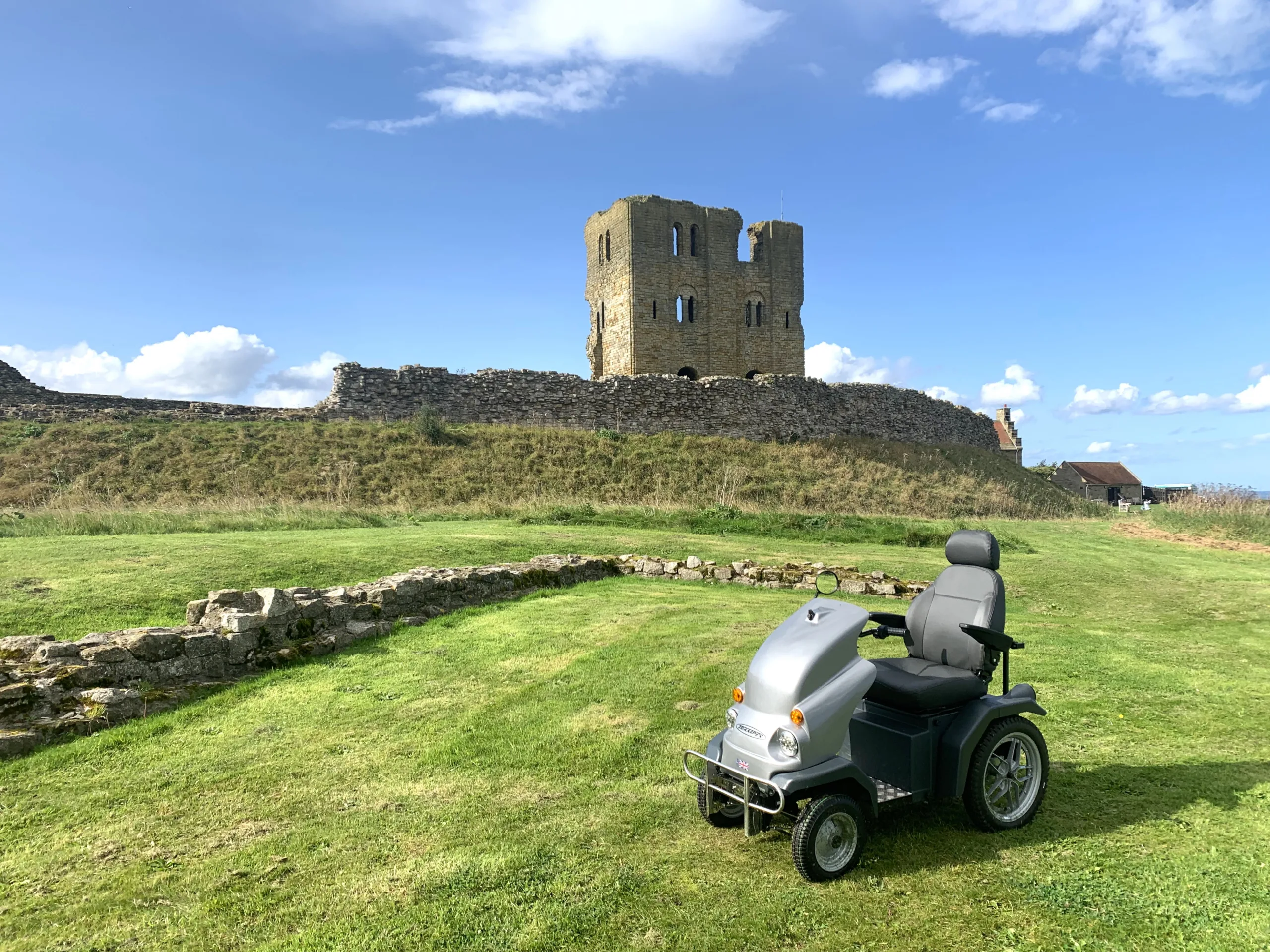 Mobility scooter parked on the grass in front of a stone-built, castle ruin on a blue sky day.