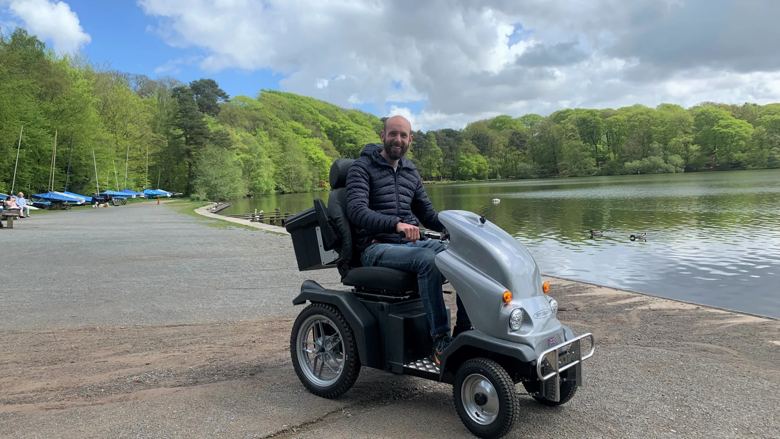 Person on a mobility scooter at the side of a body of water with lush green trees in the background. Making amazing places accessible.