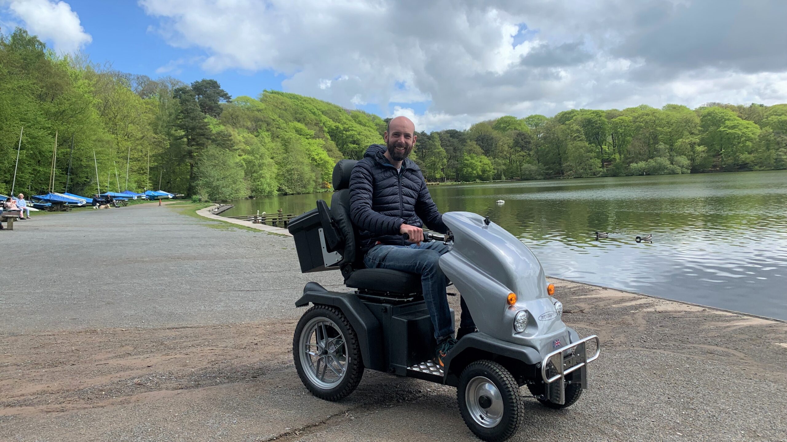 Person on a mobility scooter at the side of a body of water with lush green trees in the background. Making amazing places accessible.