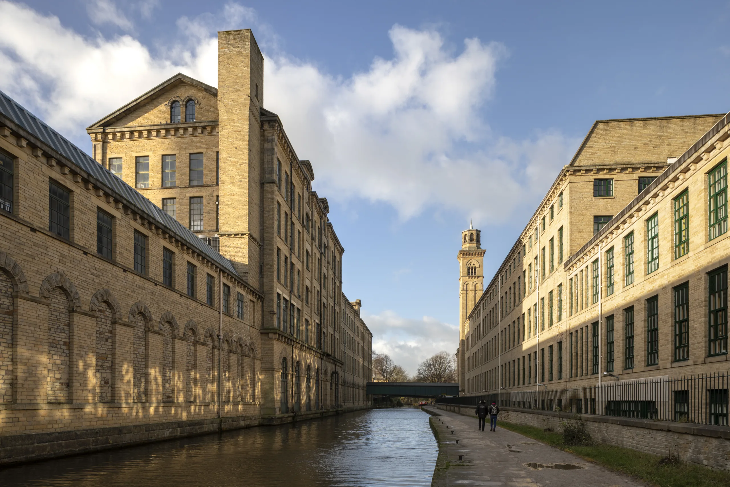 Stone built historic mill buildings stand tall at either side of a canal with reflections of the blue sky in the water.