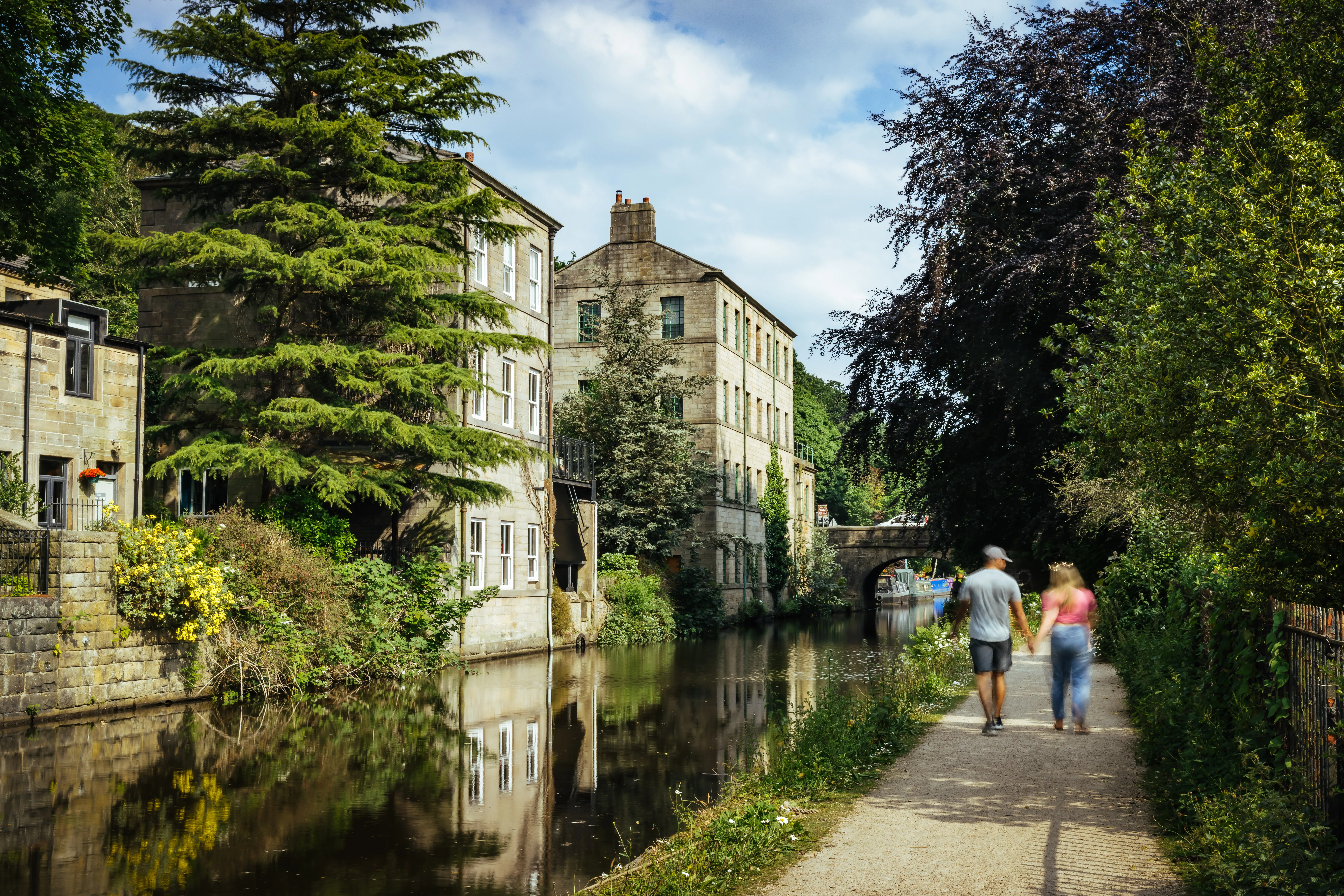 People walk along a riverside path with the river to the left and stone buildings reflected in the water.