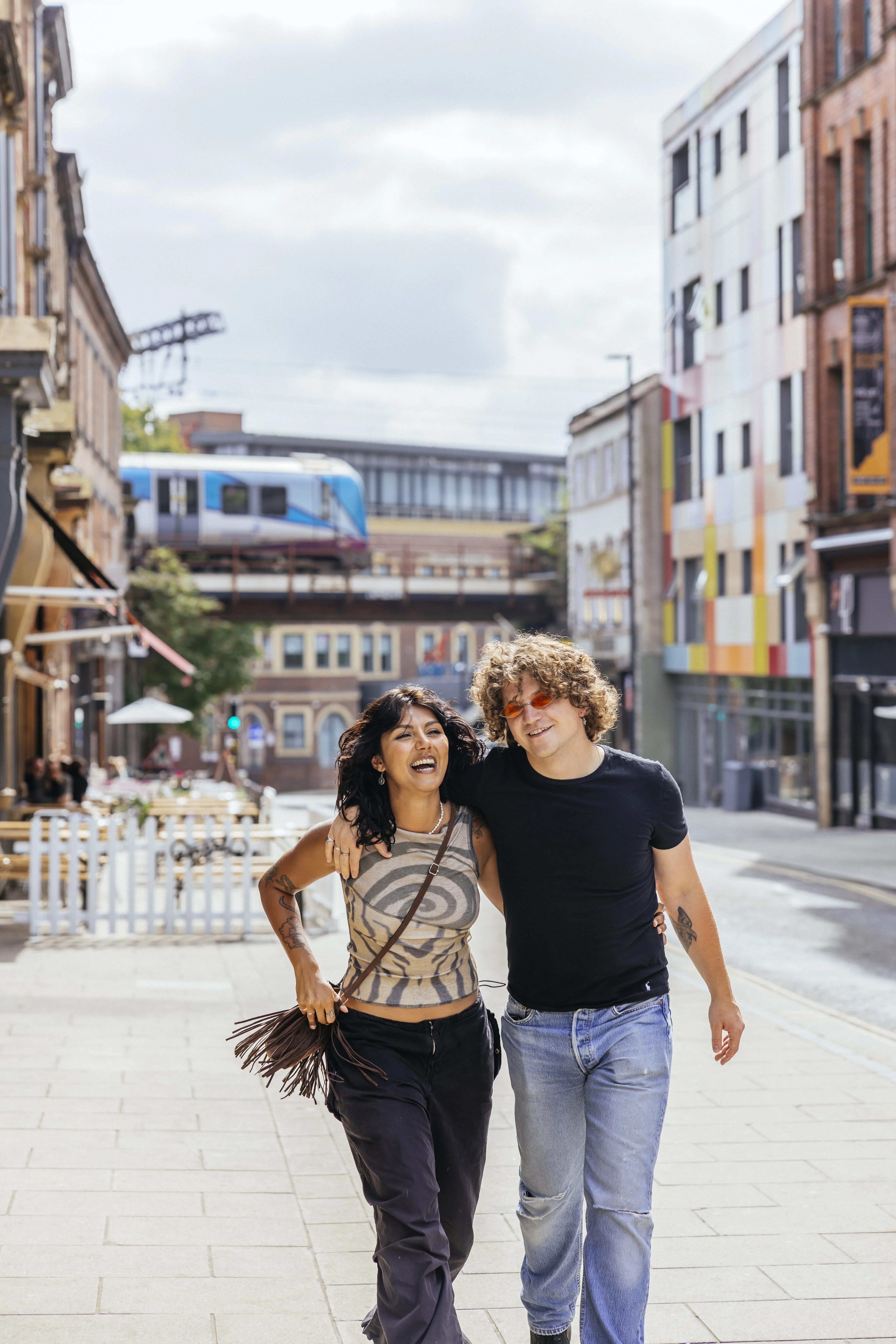 A couple walk towards the camera along a city street in Leeds. In the background stand building and a bridge with a train passing.