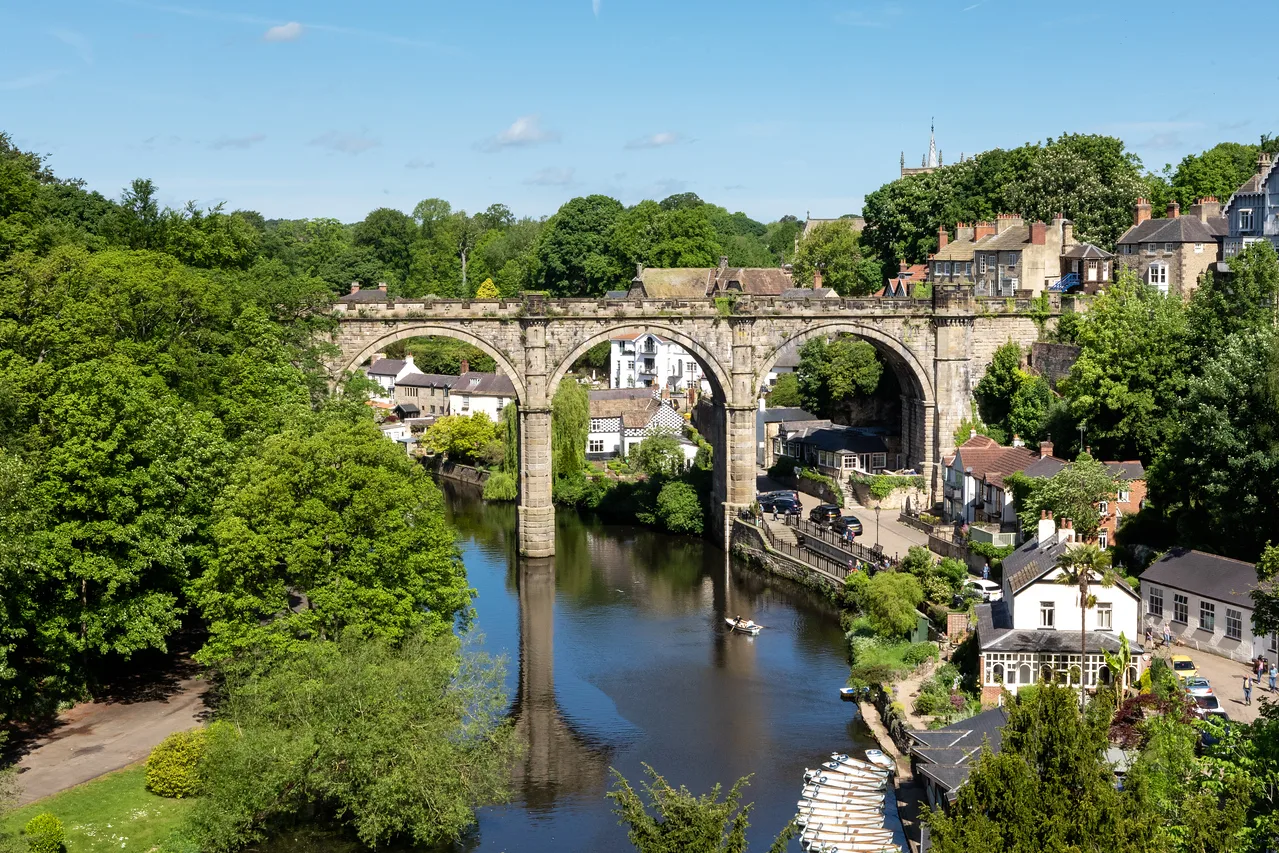 Historic stone viaduct stretches across the landscape, emerging from lush green trees towards the right of the photo where houses and building sit scattered on the hillside, some partly hidden by greenery. A river runs underneath the viaduct.