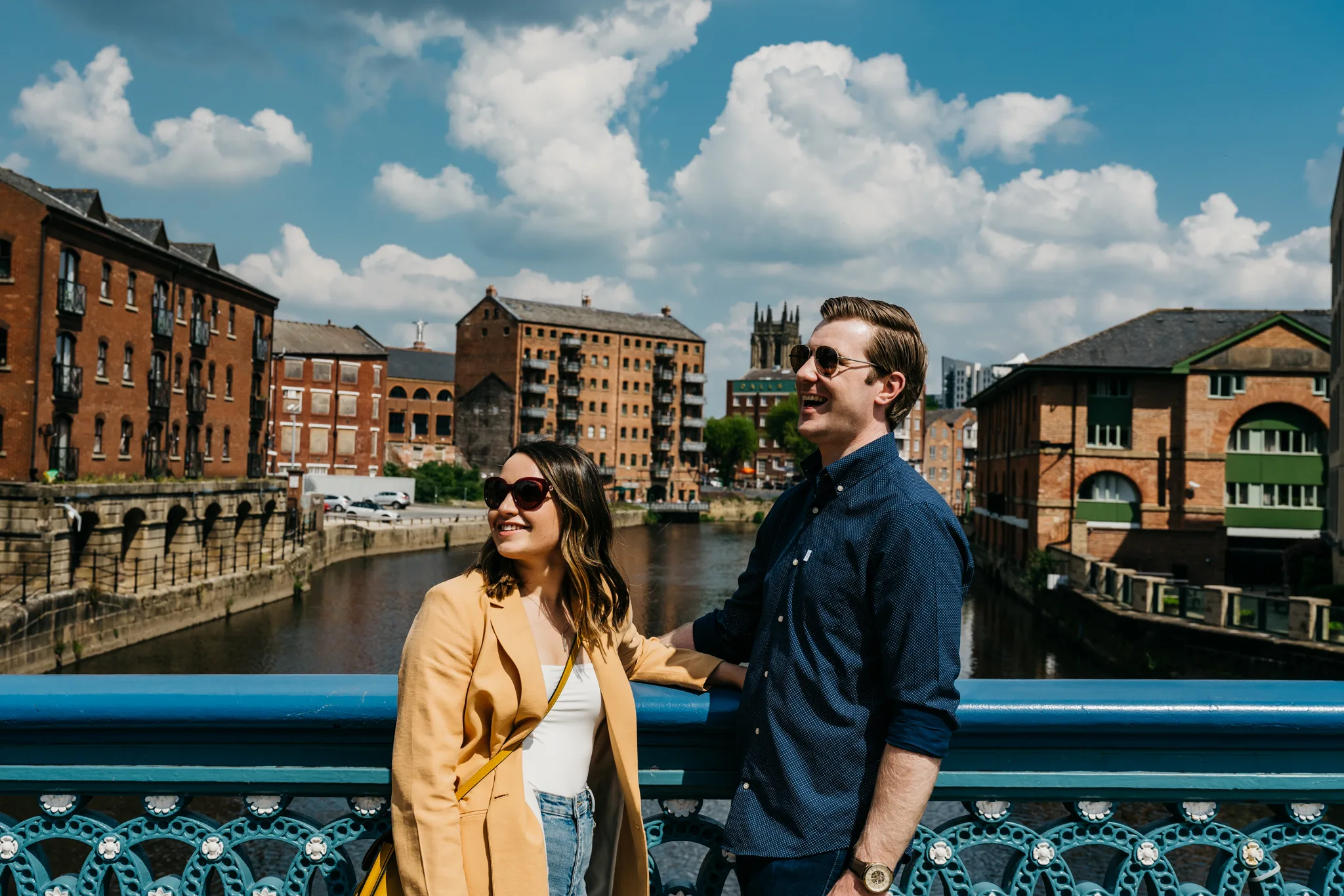 A couple stand on a bridge in the centre of Leeds smiling and looking off to the left. Behind them stands riverfront buildings, apartments and offices amid a blue sky. Leeds: A gateway to unforgettable rail adventures.