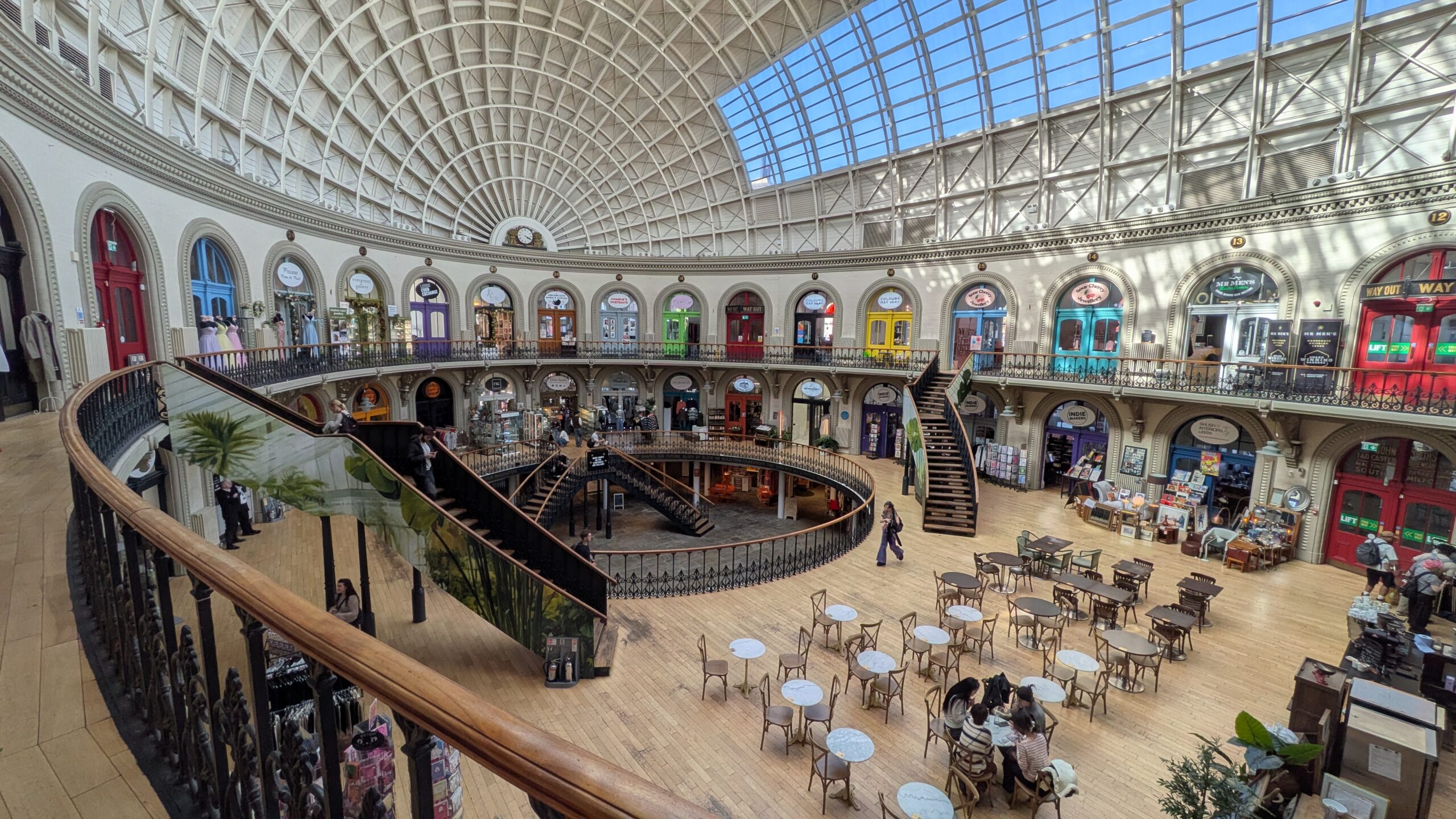 Inside view of Leeds' historic Corn Exchange with a domed roof, glass and arches. The photo looks down to the visitors exploring the centre on the lower level.