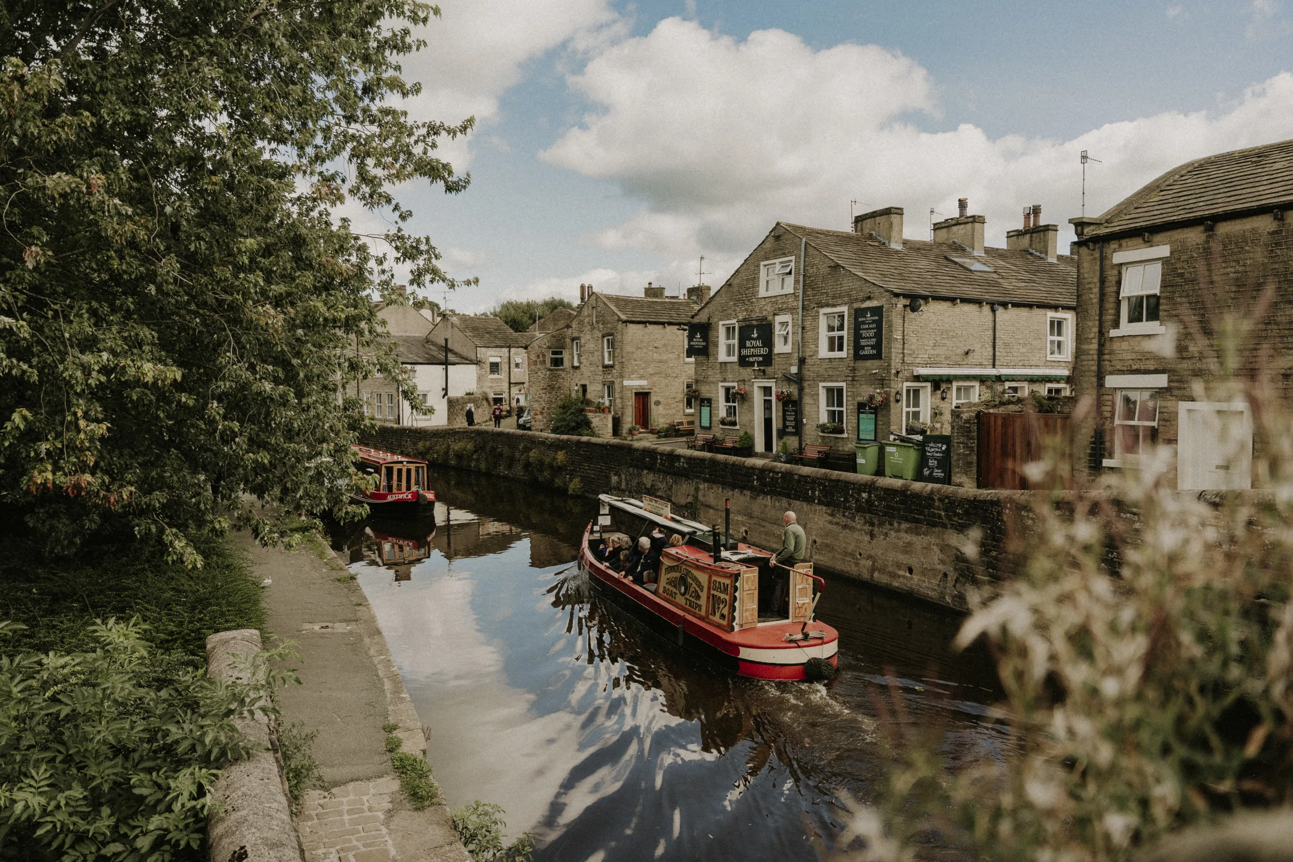 A narrowboat meanders along a calm canal passing charming buildings on the right and green foliage on the left.