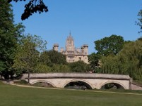A grassy field with a stone bridge stretching across the image with trees and an impressive building in the background. On a blue sky day | St. Edmund's Line