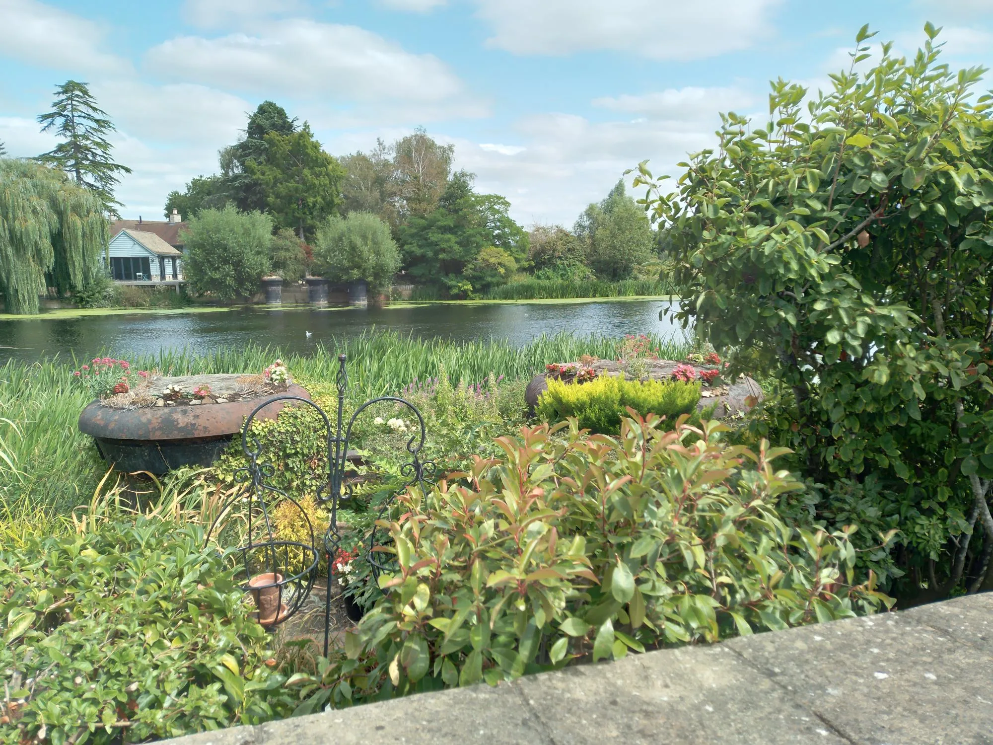 View of old bridge abutments, peeking out from green undergrowth. In the background a body of water and a residential house can be seen.