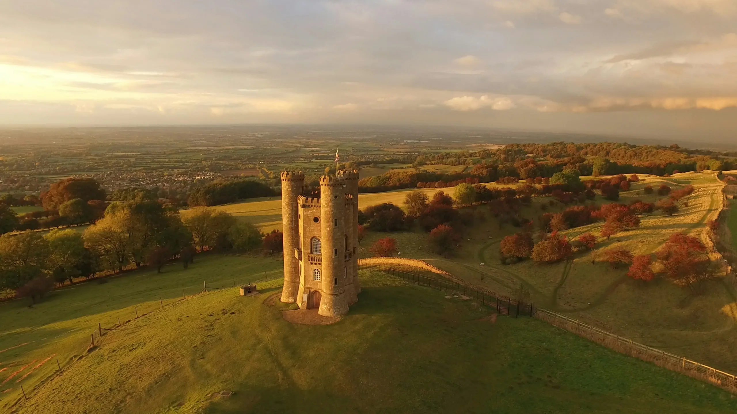 View from above of a historic tower, standing tall on top of a hill mound. Beyond the tower there is flat landscapes with trees and green fields.