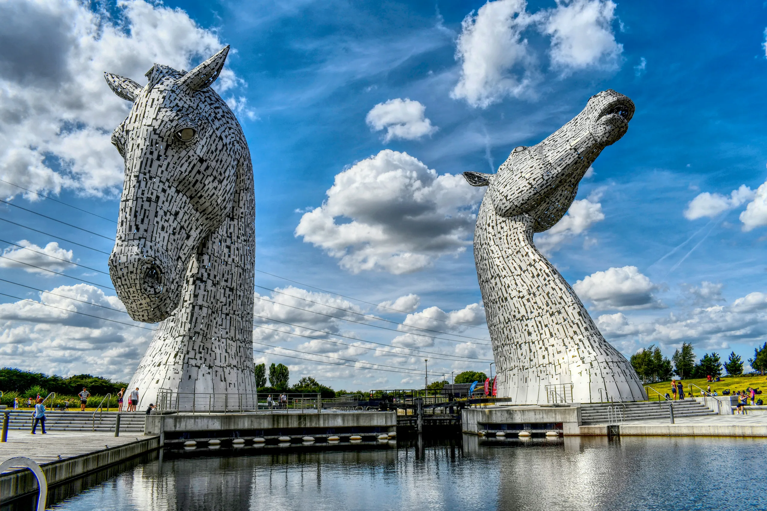 A view of two large, silver, horse head statues overlooking a body of water. The statues are reflected in the water against a blue sky with a scattering of clouds.