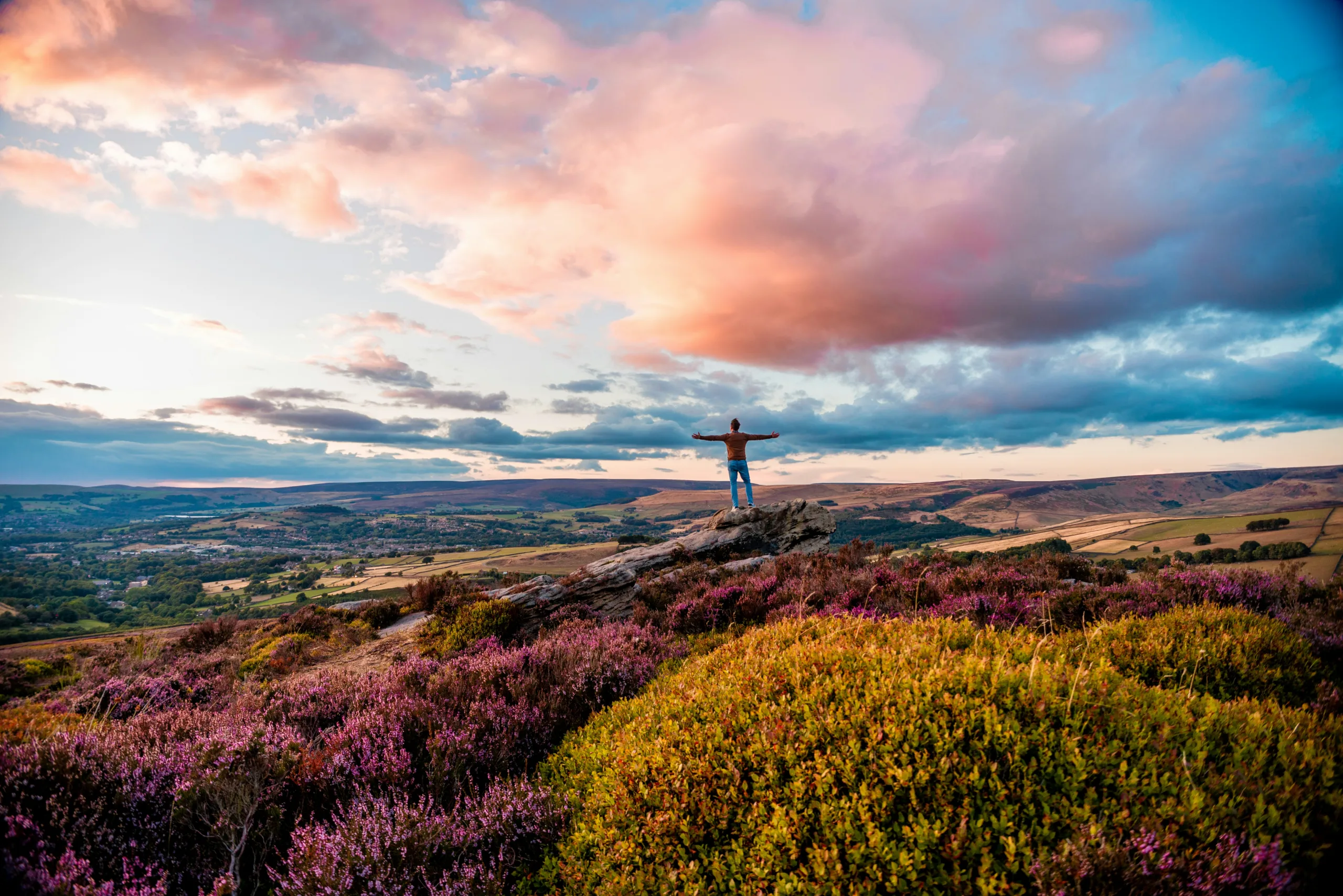 Person stood on a rocky outcrop on the top of a hill covered in moorland heath. In the valley below small houses can be seen scattered across the landscape. The sky takes up half the image with clouds and a blue sky poking through. Glossop, Discover Greater Manchester by rail